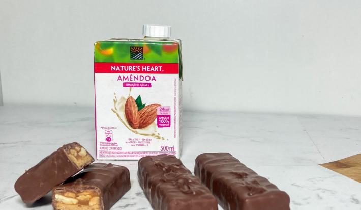 Foto em tons claros da receita de chocolate com caramelo e amendoim servida em 4 porções sobre uma mesa de mármore branca com uma embalagem de bebida vegetal de amêndoa nature's heart ao fundo