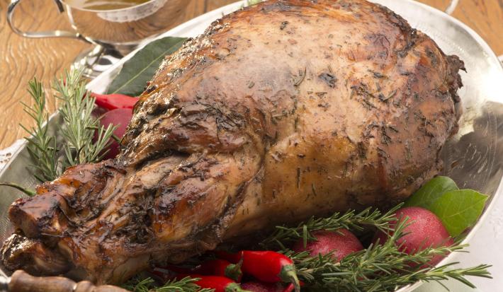 Fotografia em tons de marrom e verde, com travessa com duas alças contendo pernil assado guarnecido com ervas e pimenta vermelha, sobre dois guardanapos com um garfo, acima pratos empilhados, potinho e molheira com molhos, tudo sobre bancada de madeira.