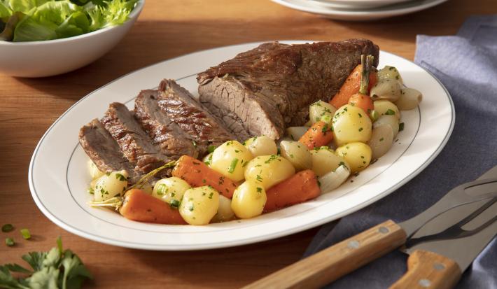 Fotografia em tons de verde em uma bancada de madeira clara, um prato oval branco com a maminha fatiada e legumes em cima dele. Ao fundo, um recipiente com salada, talheres de churrasco.