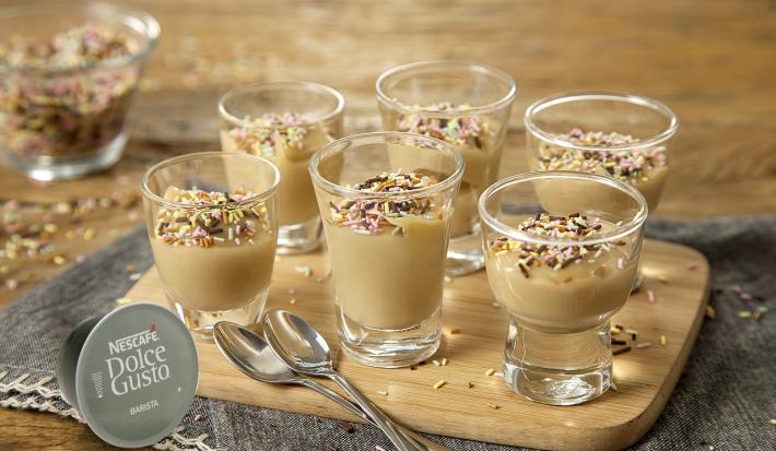 Foto de uma mesa com um pano cinza e uma tábua de madeira em cima, com 6 potinhos de vidro com brigadeiro dentro e decorados com granulado
