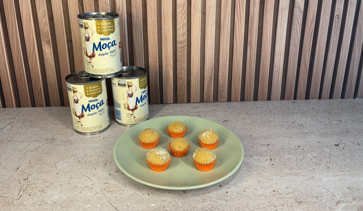Docinhos claros cobertos com açúcar em forminhas laranja, dispostos em prato verde com Leite Moça ao fundo.