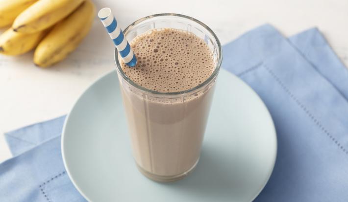 Fotografia em tons de azul e marrom de uma bancada com um paninho azul dobrado, um prato azul e um copo com a bebida de chocolate e um canudo. Ao fundo um cacho de bananas.