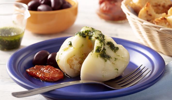 Fotografia em tons de branco em uma bancada de madeira de cor branca. Ao centro, um prato azul contendo a burrata com um garfo ao lado. Ao fundo, está uma cesta com pães e tomates secos.