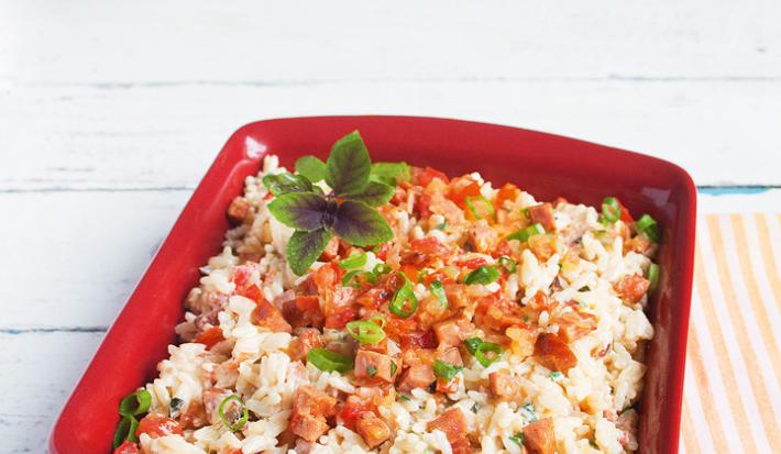 Fotografia em tons de vermelho e branco de uma bancada branca com uma pano listrado laranja e branco, uma travessa vermelha retangular com o arroz, pedaços de tomate e linguiça e manjericão.