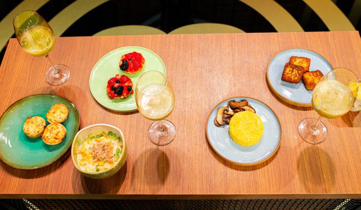 Fotografia de uma mesa de madeira com um brunch com cuscuz, quiche de alho poró, cheesecake, salada de frutas e suco de abacaxi com hortelã. As comidas estão em pratos coloridos, e o suco estão em taças, na imagem são três.