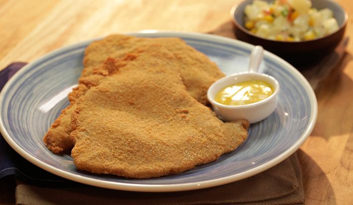 Fotografia em tons de marrom e azul de uma bancada de madeira clara, sobre ela um prato azul com dois bifes á milanesa e um mini recipiente refratário com molho.  Ao fundo um recipiente pequeno azul e redondo com vinagrete.