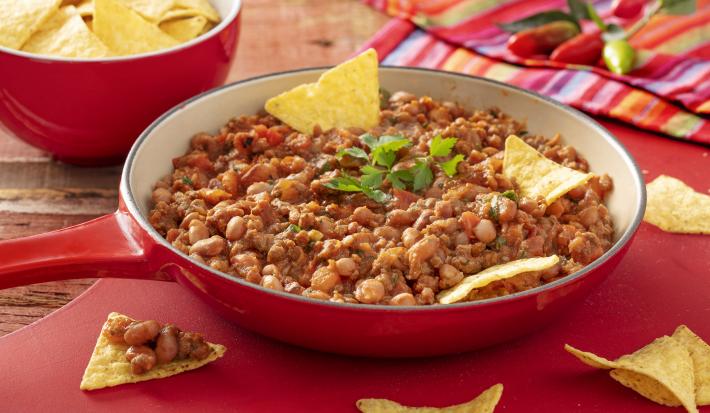 Fotografia em tons de vermelho em uma bancada de madeira escura, um jogo americano vermelho, um pano colorido, uma frigideira grande vermelho com o feijão mexicano dentro. Ao fundo, um pote vermelho com nachos.