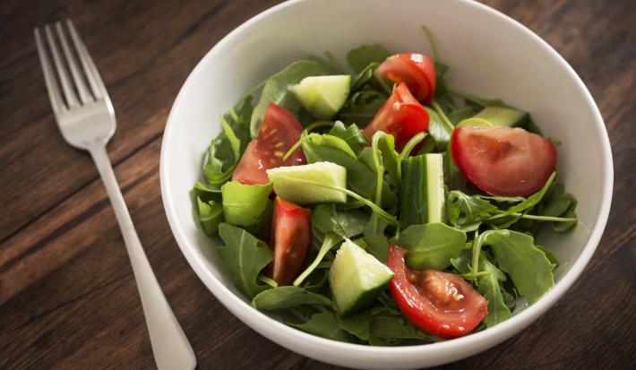 Fotografia de um recipiente fundo branco com uma salada de folhas, pepino e tomates. Ao lado do recipiente, um garfo, que está sobre uma mesa de madeira escura.