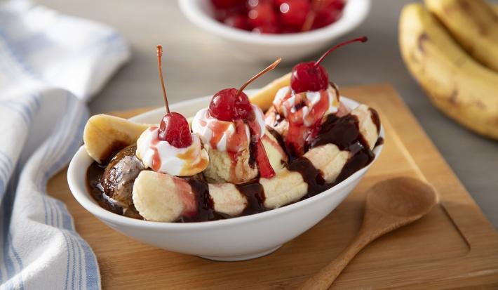 Fotografia em tons de vermelho em uma bancada de madeira de cor cinza. Ao centro, uma tábua de madeira com uma tigela em cima contendo o banana split. Ao lado, há uma colher de madeira e um pano listrado azul. Ao fundo, 2 bananas e algumas cerejas.