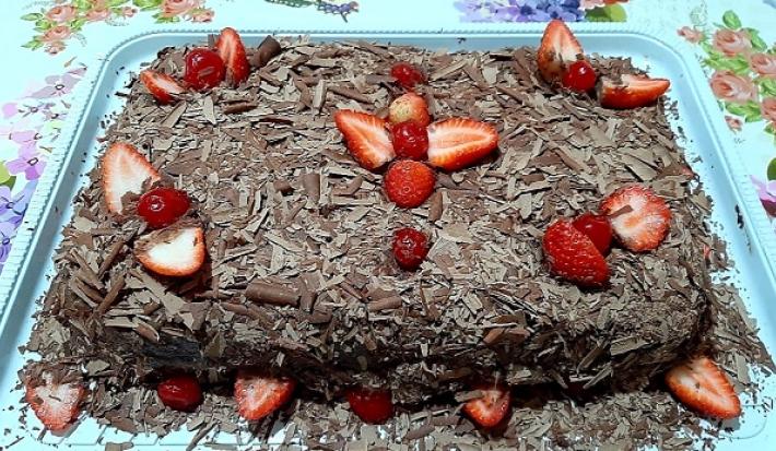 Foto em tons de marrom da receita de bolo de chocolate com morangos servida inteira e decorada com morangos e cerejas em cima