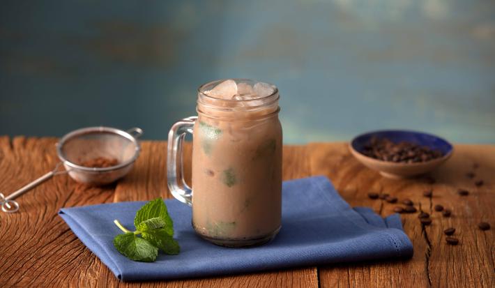 Fotografia em tons de azul em uma bancada de madeira escura, um pano azul, uma caneca alta de vidro com o cappuccino gelado de cacau, banana e hortelã. Ao lado, uma peneira com o cacau e um potinho com algumas gotas de chocolate.