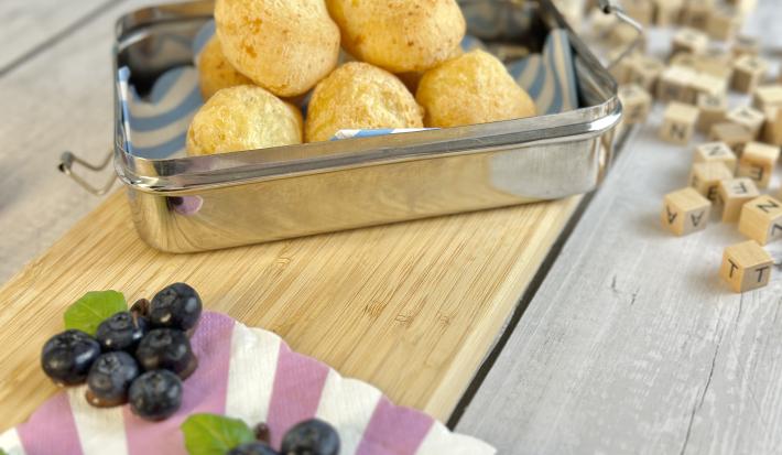 Fotografia em tons de roxo em uma bancada de madeira cinza com uma tábua de madeira marrom, uma lancheira em cima com pão de queijo feito de tapioca dentro dela. Ao lado, objetos e brinquedos infantis.
