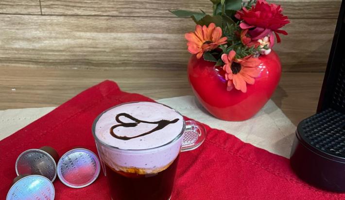 Fotografia em tons de vermelho em uma bancada de madeira, um pano vermelho e uma xícara com café e espuma rosa decorado com um coração com calda de chocolate ao centro. Ao fundo, um vaso vermelho com flores e em formato de coração.