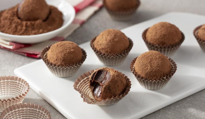 Fotografia em tons de branco, cinza e marrom de uma bancada cinza com um prato retangular branco com trufas de chocolate zero em forminhas marrom. Ao fundo um paninho xadrez branco e vermelho, um recipiente branco redondo com chocolate em pó e uma trufa.