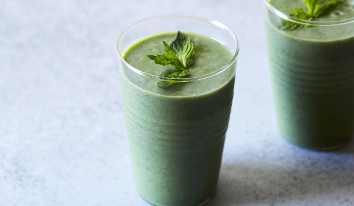 Fotografia de dois copos de vidro com um refresco de hortelã, e por cima, folhas de hortelã. As vitaminas estão sobre uma mesa em tons de azul claro.