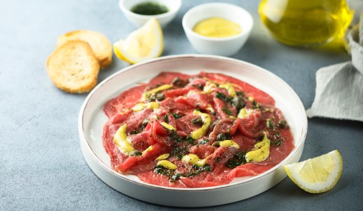 Foto da receita de Carpaccio com Alcaparras e Molho de Mostarda. Observa-se um prato branco grande com a carne fina dentro em rodelas, com molho e alcaparras.