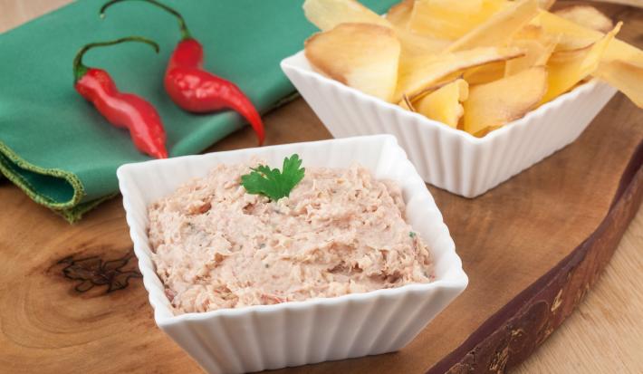 Fotografia em tons de branco, verde e marrom, com dois potinhos brancos contendo patê na cor rosa e mandioca chips, ao lado guardanapo verde com duas pimentas vermelhas, tudo sobre peça de madeira maciça.