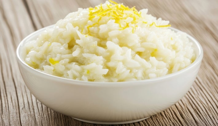 Em cima de uma mesa, tigela redonda branca com porção de arroz cremoso, decorado com raspas de limão.