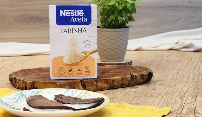 Fotografia em tons de marrom com um prato redondo ao centro. Em cima do prato existe duas panquecas de chocolate com aveia recheadas com um creme de iogurte. Ao fundo existe uma embalagem de Farinha de aveia NESTLÉ