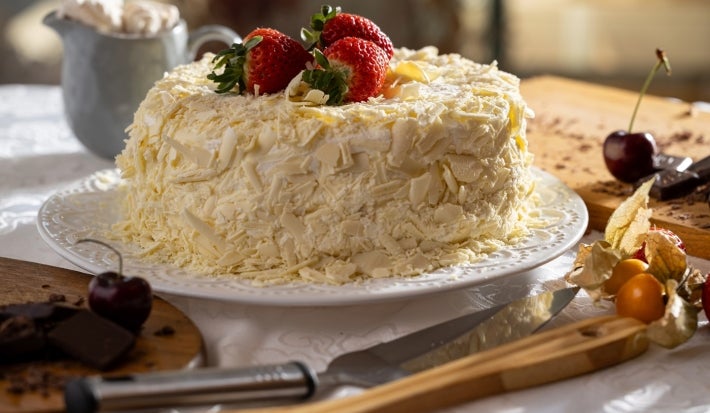 O mesmo bolo aparece inteiro, coberto com raspas de chocolate branco e decorado com morangos no topo. Está sobre um prato branco, cercado por utensílios e frutas, pronto para ser servido em uma mesa posta.