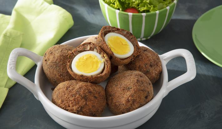 Foto da receita de Bolovo. Observa-se um recipiente com 6 bolovos, sendo que um está cortado no meio. Atrás, um recipiente com salada decora.