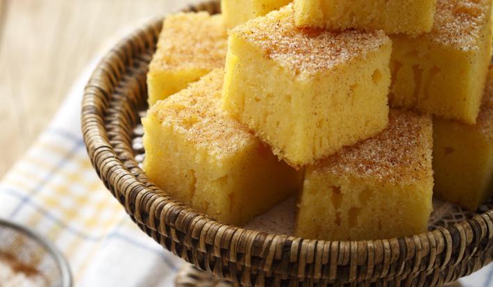 Foto de pedaços quadrados de bolo de fubá em um recipiente marrom em cima de um pano branco com detalhes laranja e azul