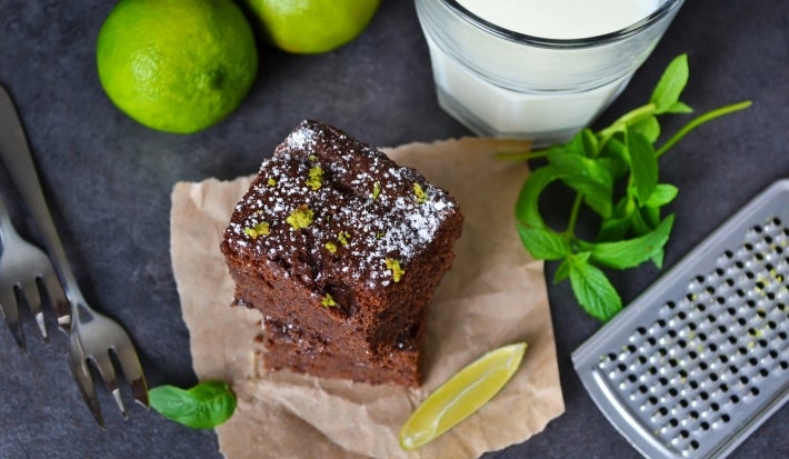 Pedaços de bolo de chocolate empilhados sobre papel manteiga, decorados com açúcar de confeiteiro e raspas de limão. Limas inteiras e uma fatia de limão ao lado, com um copo de leite e utensílios, sugerindo um momento de degustação.