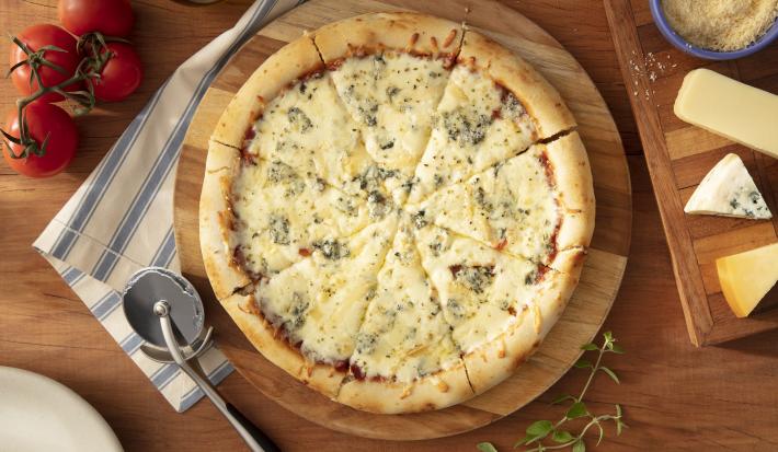 Imagem vista de cima de uma pizza redonda de 4 queijos. Ao lado uma tábua com queijos e um cortador de pizza.