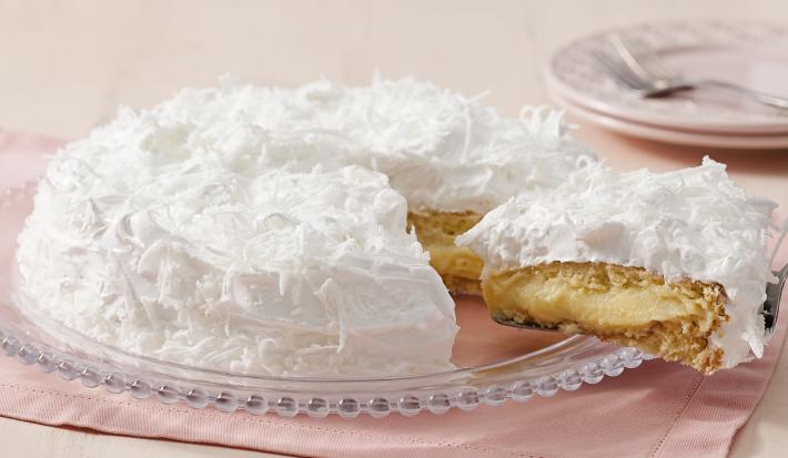 Fotografia de um bolo coberto com marshmallow branco e raspada de coco em cima. Dentro do bolo creme e massa brancos. O prato está em cima de um guardanapo de papel rosa claro.
