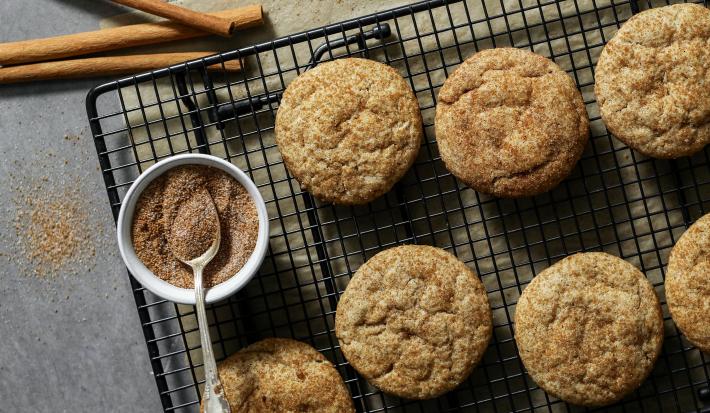 Fotografia vista de cima de sete cookies com açúcar e canela sobre uma grade preta. Ao lado dos cookies e por cima da grade, tem um pequeno pote de vidro branco com açúcar e canela, e uma colher de chá apoiada sobre ele.
