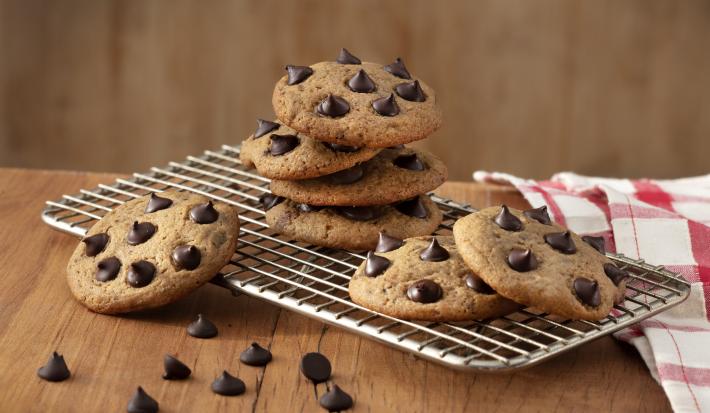 Fotografia em tons de marrom em uma bancada de madeira, um pano xadrez vermelho, um suporte de grelha com alguns cookies com gotas de chocolate em cima dele.