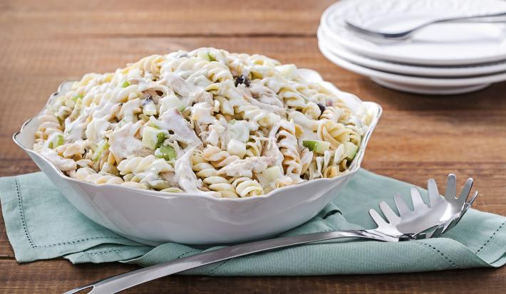 Fotografia em tons de azul em uma bancada de madeira escura, um pano azul com um recipiente redondo branco em cima dele e a salada de macarrão com peito de frango dentro dele. Ao lado, uma colher de servir macarrão.