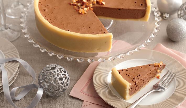 fotografia em tons de cinza e marrom de uma bancada cinza, contém um suporte para servir sobremesa transparente e redondo com uma torta com mousse de chocolate. Ao lado um prato rendo e bege com uma fatia da torta e um garfo ao lado para servir. fotografia em tons de cinza e marrom de uma bancada cinza, contém um suporte para servir sobremesa transparente e redondo com uma torta com mousse de chocolate. Ao lado um prato rendo e bege com uma fatia da torta e um garfo ao lado para servir.