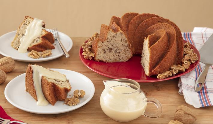 Foto da Receita de Bolo de Nozes com Molho doce. Observa-se dois pratos de sobremesa com pedaços do bolo e a calda. Ao lado direito, um prato redondo vermelho grande com o restante do bolo, decorado por nozes.