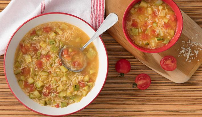 Fotografia em tons de branco, vermelho e marrom, sopeira branca com borda vermelha contendo sopa com legumes, arroz e uma concha, ladeada por guardanapo branco e vermelho, ao lado pote com sopa sobre tábua, tomate, tudo sobre bancada em tons de marrom.