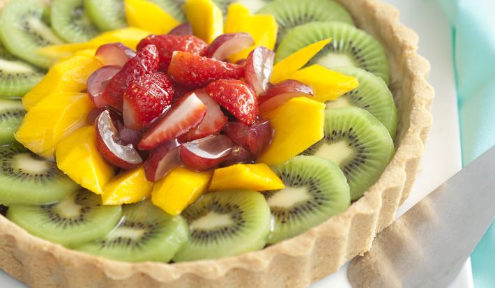 Fotografia em tons de verde, branco, amarelo e vermelho, com torta de frutas sobre porcelana branca com uma espátula e guardanapo verde ao lado, tudo sobre bancada em tons de branco e cinza.