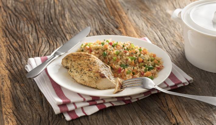 Fotografia em tons de marrom com um pano branco com listras vinho e um prato branco raso com o arroz feito na panela de pressão com legumes e um filé de frango.