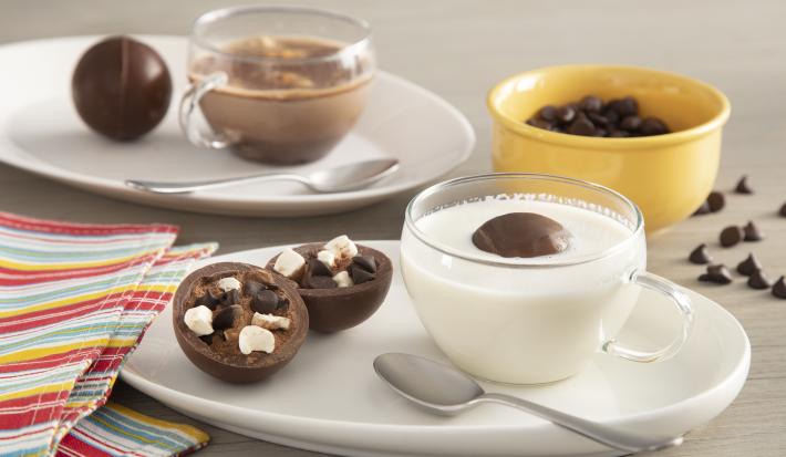 Fotografia em tons de branco e amarelo de uma bancada cinza, ao centro um prato com uma xícara com leite e o chocobomb e ao lado um chocobomb aberto, mostrando a camada de chocolate e o recheio.