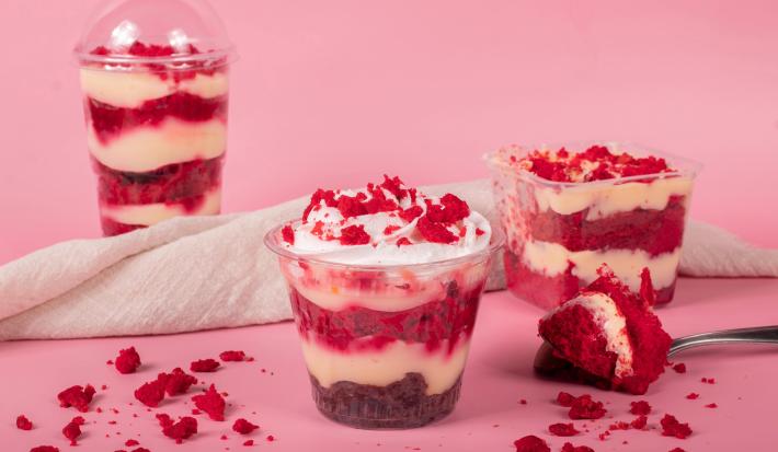 Fotografia em tons de rosa com alguns bolos de pote de red velvet ao centro. Dentro de cada pote, existe camadas de creme branco alternadas com bolo red velvet