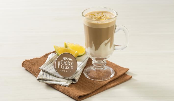fotografia em tons de branco e marrom tirada de frente, contém um pano marrom com uma xicara transparente com a bebida de NESCAFÉ Dolce Gusto Café Au Lait com NESTLÉ Creme de Leite. Ao lado uma cápsula de Dolce Gusto e ao fundo fatias de laranja