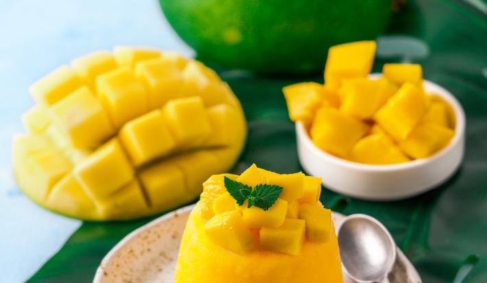 Fotografia de um flan de manga e calda de carambola, com pequenos pedaços de manga por cima. Ao lado da sobremesa, uma colher pequena que está sobre um miniprato de cerâmica branco com pontos pretos. Ao fundo, manga em pedaços e uma inteira.