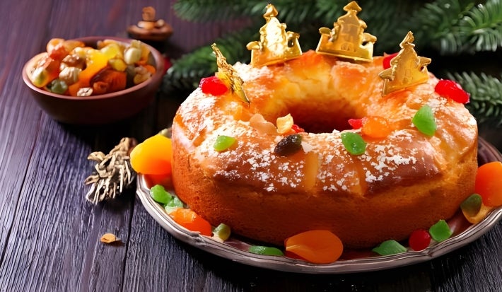Fotografia de um bolo festivo, decorado com frutas cristalizadas coloridas e coroas douradas, apresentado em um prato prateado.