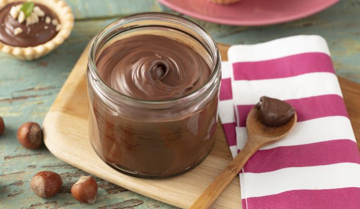 Fotografia em tons de roxo em uma bancada de madeira esverdeada, um pano branco com listras roxas, uma tábua de madeira, um potinho cilíndrico de vidro com o creme de avelã dentro dele. Ao fundo, mini tortinhas com o creme e avelãs espalhadas na mesa.