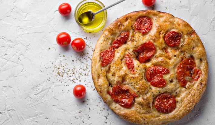 Fotografia vista de cima de uma focaccia com tomate cereja assado, sal grosso e azeite. Ao lado da focaccia tem um recipiente de vidro pequeno com azeite, ao lado alguns tomates cereja, sobre uma mesa branca com texturas.