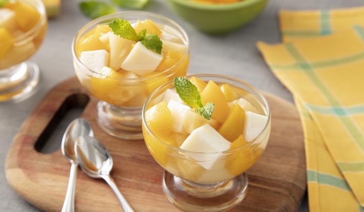 Foto da receita de Gelatina de Verão. Observa-se três potes individuais de vidro com as gelatina em quadrados e frutas amarelas picadas sobre uma tábua de madeira. Ao lado dos potes, duas colherinhas acompanham.
