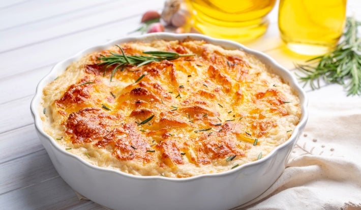 Em cima de uma mesa, refratário com batata gratinada, decorado com cebolinha alecrim.