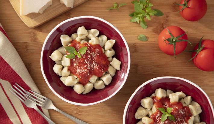 Imagem vista de cima de dois bowls vermelhos com nhoque de mandioca e molho de tomate. Ao lado, tomates, manjericão, garfos e queijo parmesão.