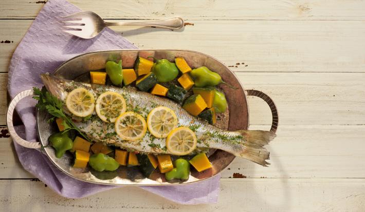 Fotografia em tons de lilás em uma bancada de madeira clara com um pano lilás, uma panela de ferro com o peixe assado com legumes dentro dela.