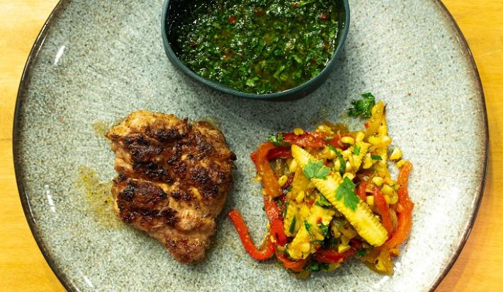 Fotografia em tons de amarelo com um prato azul ao centro. Em cima do prato existe uma receita de churrasco de timo acompanhado de chimichurri e pimentões e mini-milhos tostados.