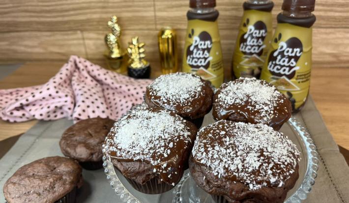 Fotografia em tons de marrom e dourado com um recipiente de vidro ao centro. Em cima do recipiente existe vários muffins cobertos com chocolate e coco ralado. Ao fundo existe três garrafas de PANTÁSTICA, uma mistura pronta para panquecas.